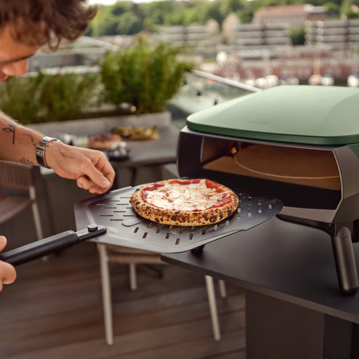 Person placing a pizza using Witt Soft Grip Pizza Peel Black on a portable outdoor oven on a balcony.