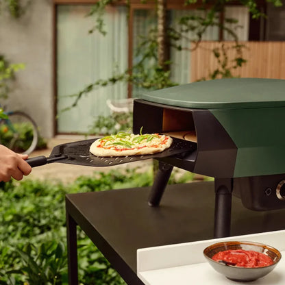 Person placing a pizza onto an outdoor pizza oven in a garden setting.