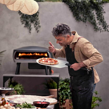 Man holding a plate of food in front of an outdoor grill with plants and a gray wall in the background.