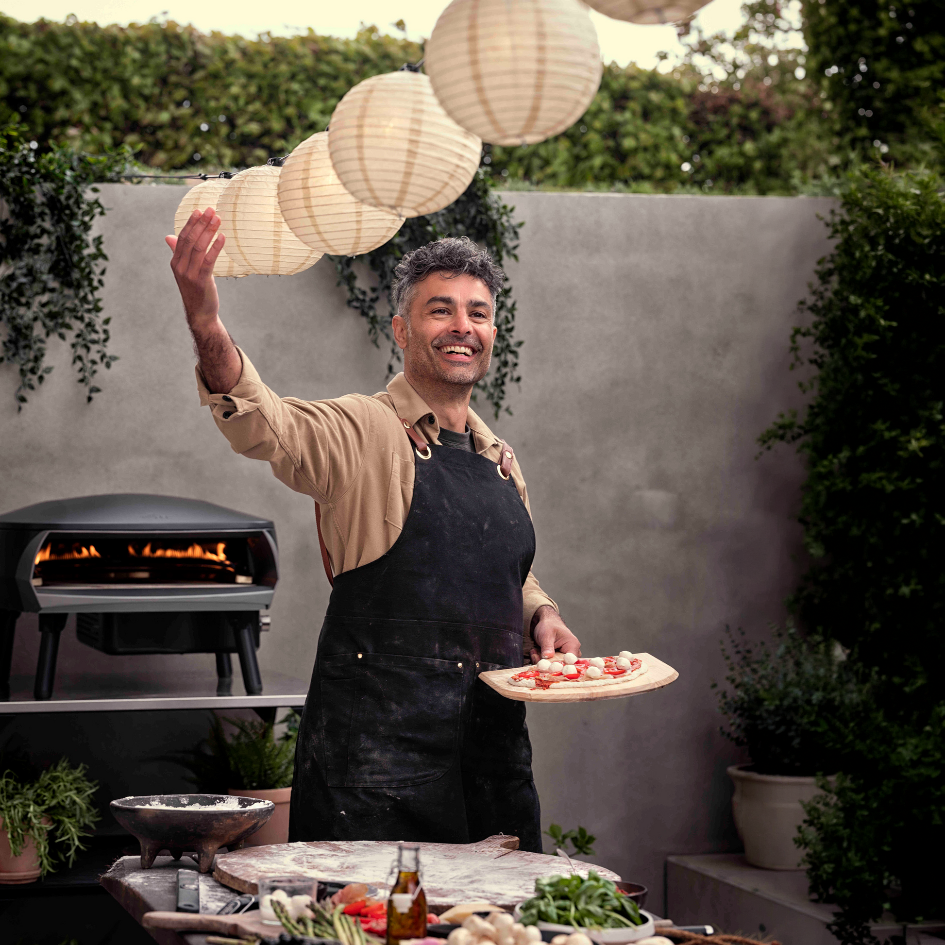 Man holding a pizza in an outdoor setting with a grill and lanterns.