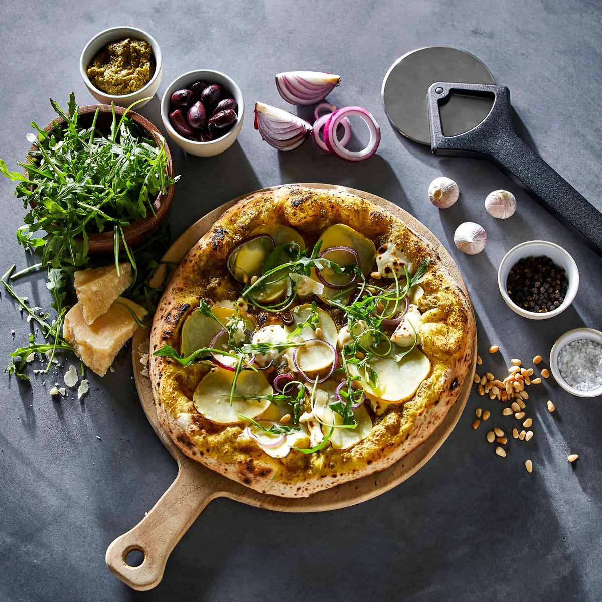 Vegetable pizza on a wooden board with ingredients and a Witt black pizza cutter on a dark surface