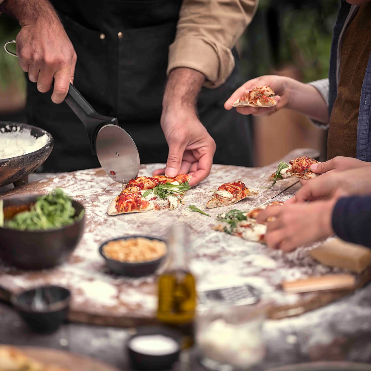 People preparing and enjoying pizza at a table with various ingredients and a Witt Witt Pizza Cutter Wheel Stainless Steel.
