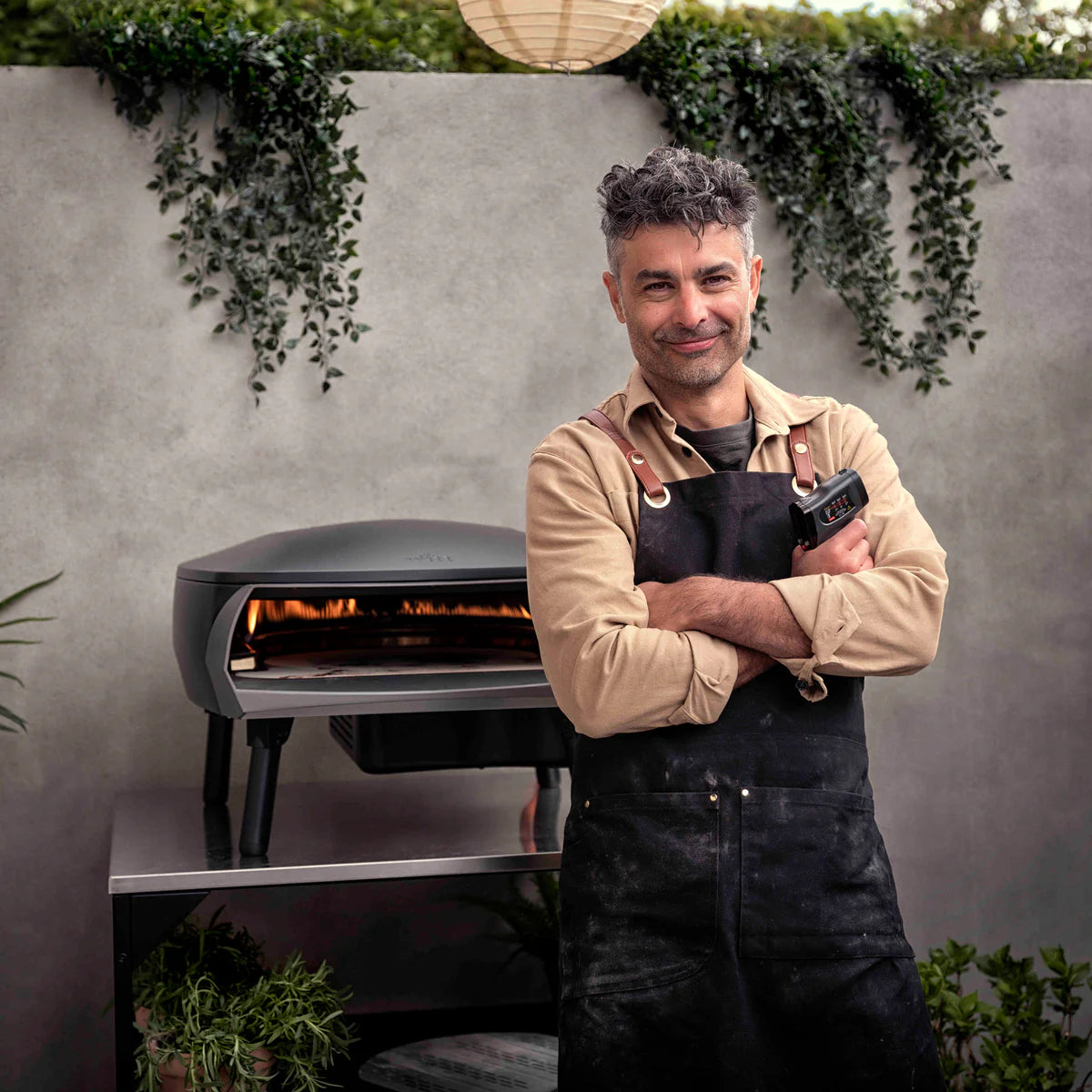 Man in an apron standing next to a Witt pizza oven outdoors with greenery in the background. He is holding a Witt Pizza Infrared Temperature Gun Black