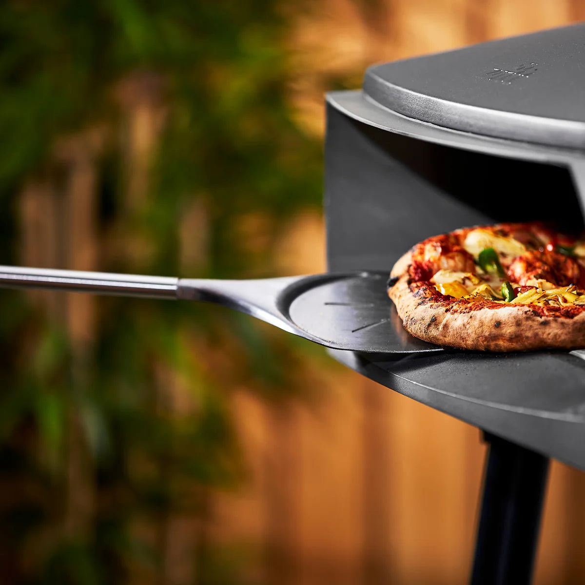 Portable pizza oven with a pizza being cooked outdoors, using Witt turning peel to takeout the pizza from oven