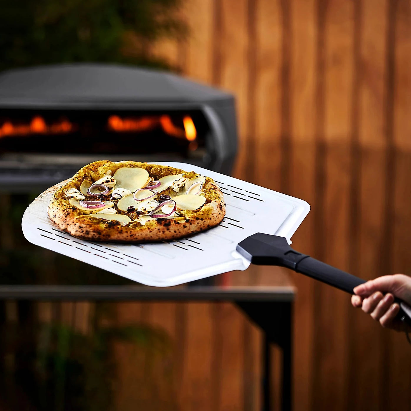 Person using a pizza peel to take a pizza out of an outdoor pizza oven.