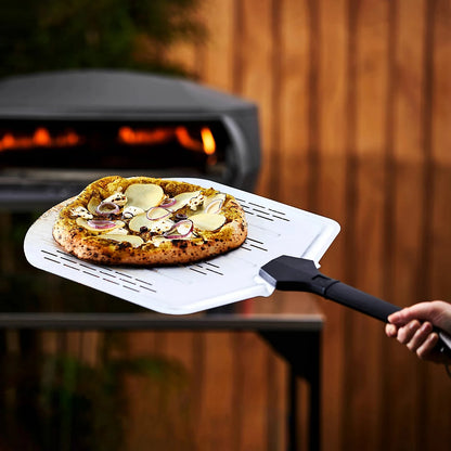 Person using a pizza peel to take a pizza out of an outdoor pizza oven.